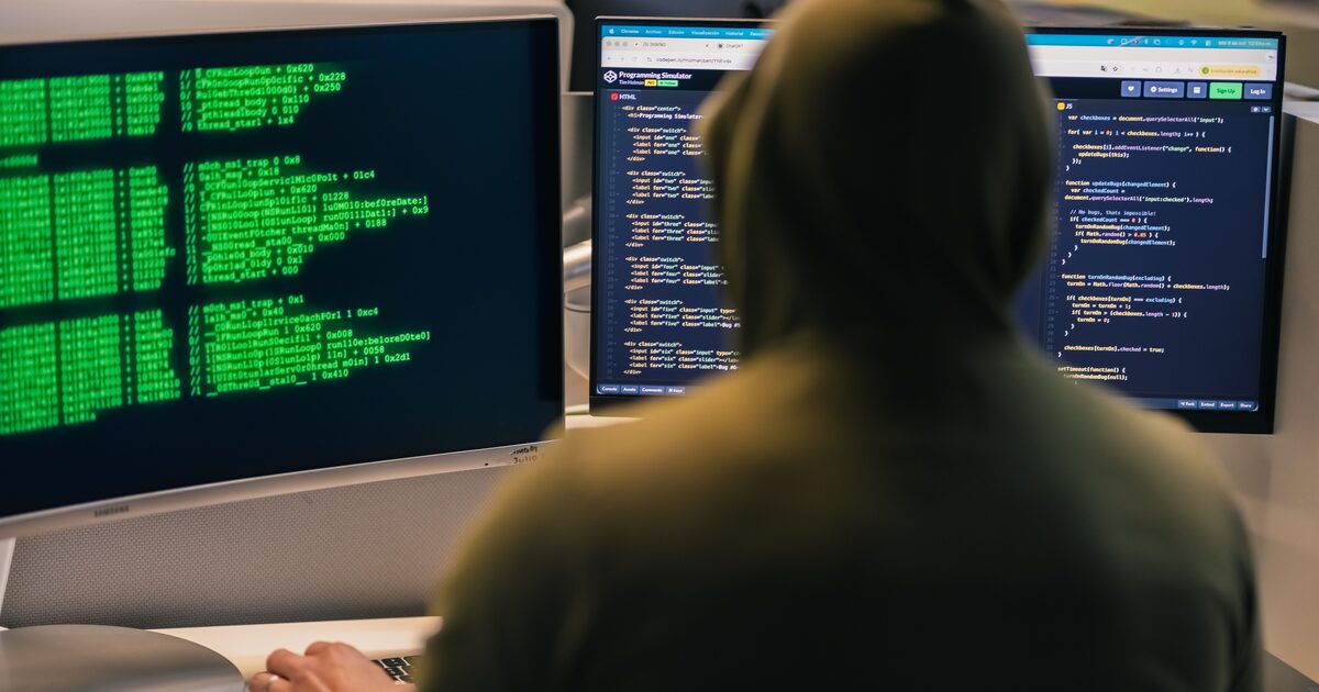Person i huvtröja sedd bakifrån framför tre datorskärmar med programkod